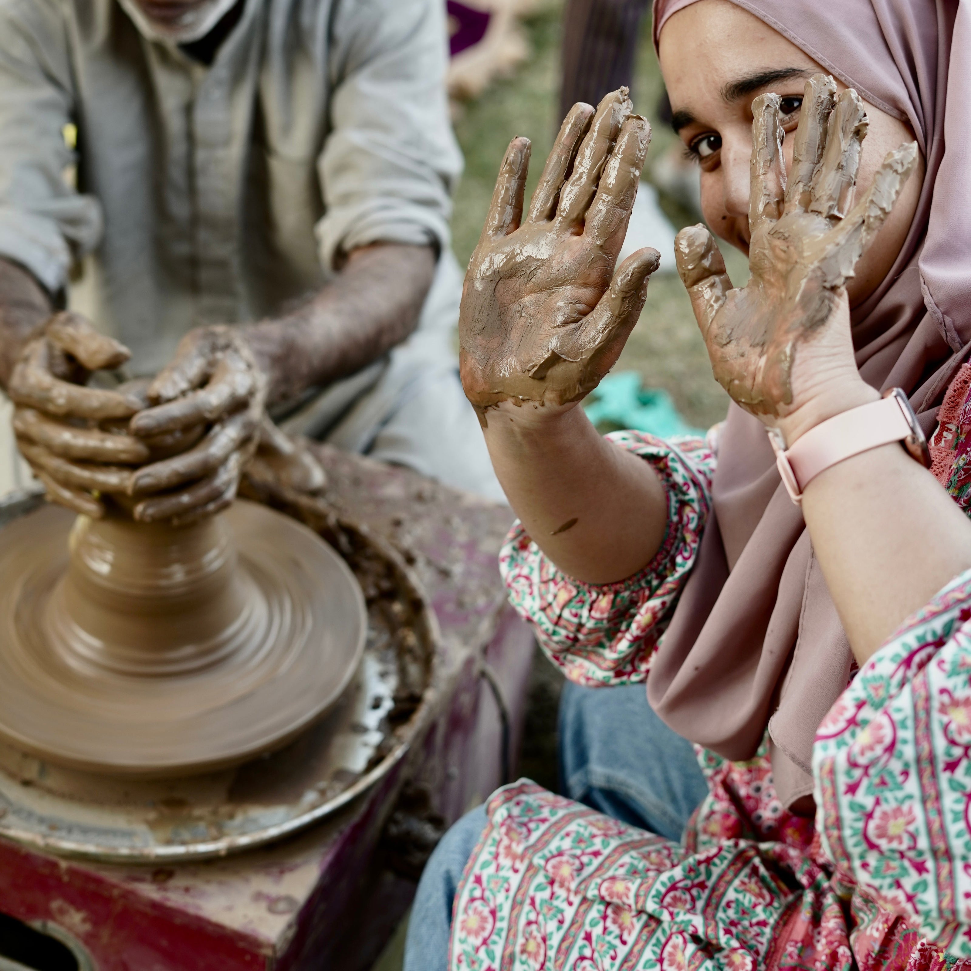 Two people working with clay on a pottery wheel outdoors.