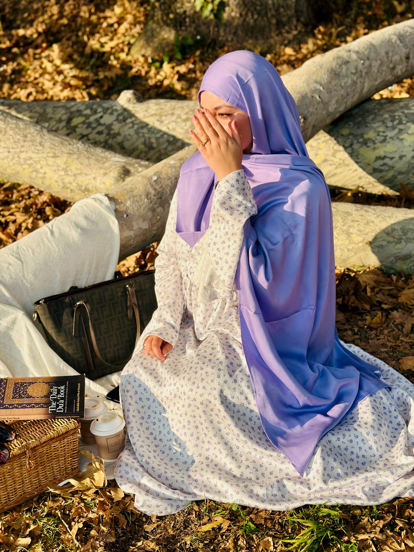 Person wearing a purple hijab and white dress sitting outdoors with a basket and book.