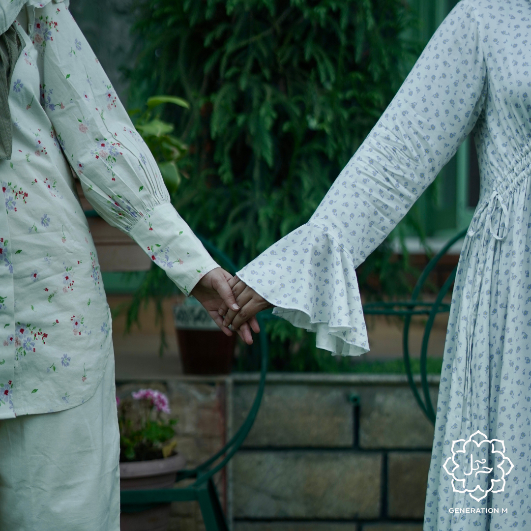Two people holding hands with a blurred background of greenery and a stone wall.